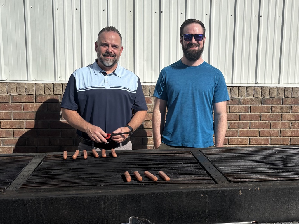 Two men stand at an outdoor BBQ grilling hotdogs