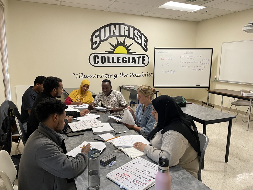 Adults gathered around a table collaborating with the Sunrise Collegiate logo in the back