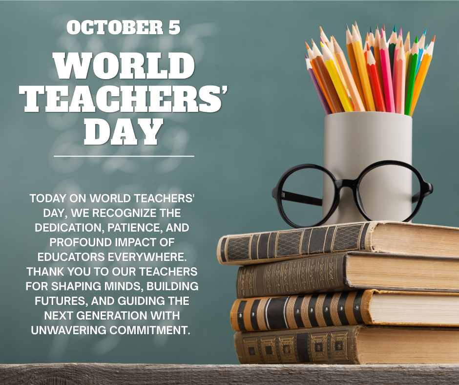 A stack of books sits on a desk in front of a chalkboard with glasses and a cup full of coloured pencils