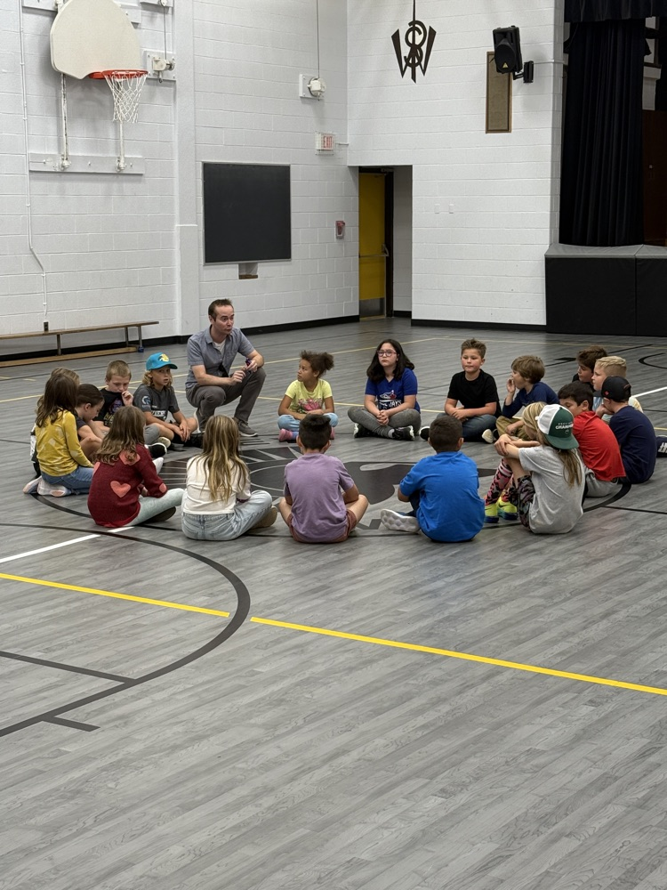 a class of students sitting in a circle in the gym listening to instructions from the teacher 