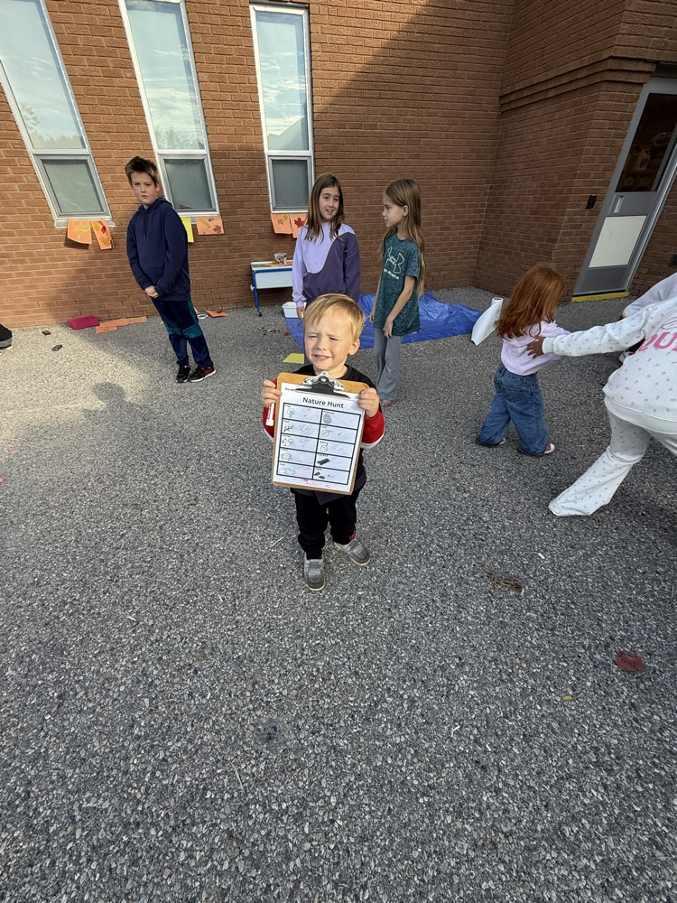 One kindergarten student who is on a nature hunt holding a clipboard smiling at the camera