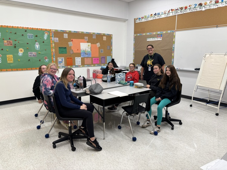 8 Grade 8 girls sitting at a table and their teacher standing beside them. Student Action Committee