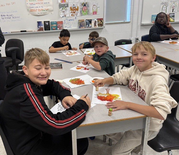 Some older students sitting at a desk, smiling at a camera while they are painting, fall art