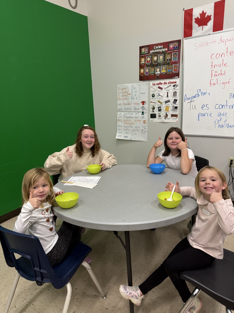 A group of student students sitting at a round table, smiling at the camera while they’re eating homemade applesauce