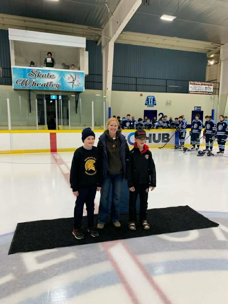 Two young students standing on either side of their teacher, Mrs. Hopper at centre ice for the hockey game