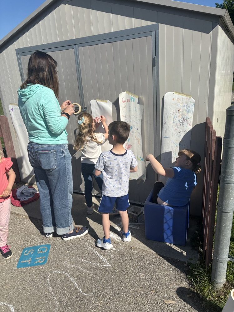 students asking to write outdoors. teacher putting up paper on the shed so they can write 