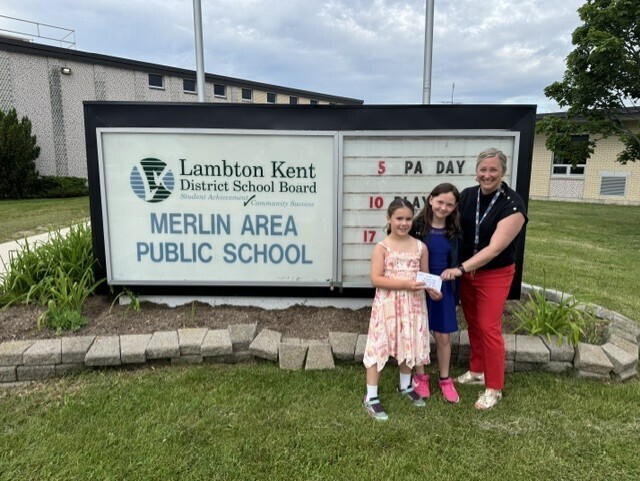 Annabelle F and Ava S present their $112.17 lemonade stand donation to Principal Harmony Queen Munroe on June 9, 2026, in support of the Merlin Area Public School playground fundraising campaign