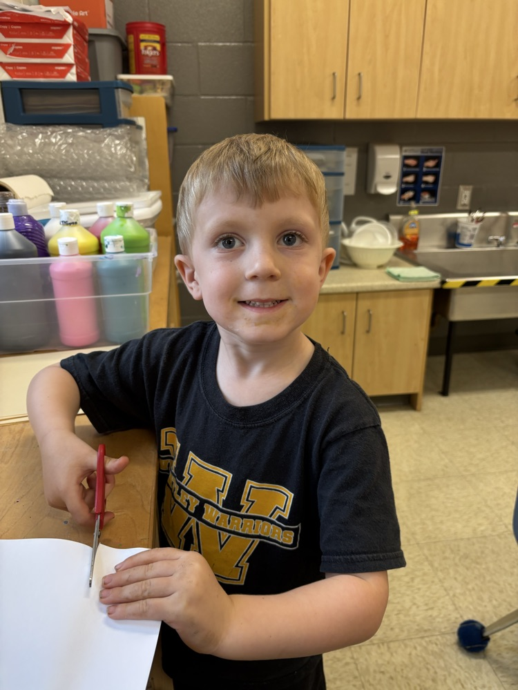 Young student wearing his Warrior shirt and smiling at the camera while cutting a piece of paper