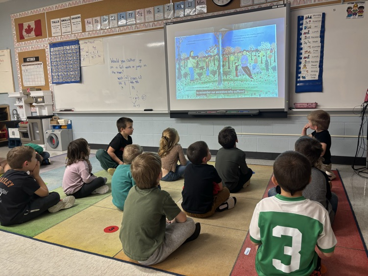 classroom of students sitting on a carpet listening to a story on the Smartboard