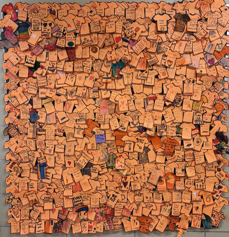 A bulletin board filled with small, paper, orange shirts created by students.