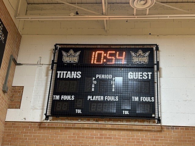 new scoreboard mounted on the wall in the TDHS gym, white and brown brick background with black lighted scoreboard