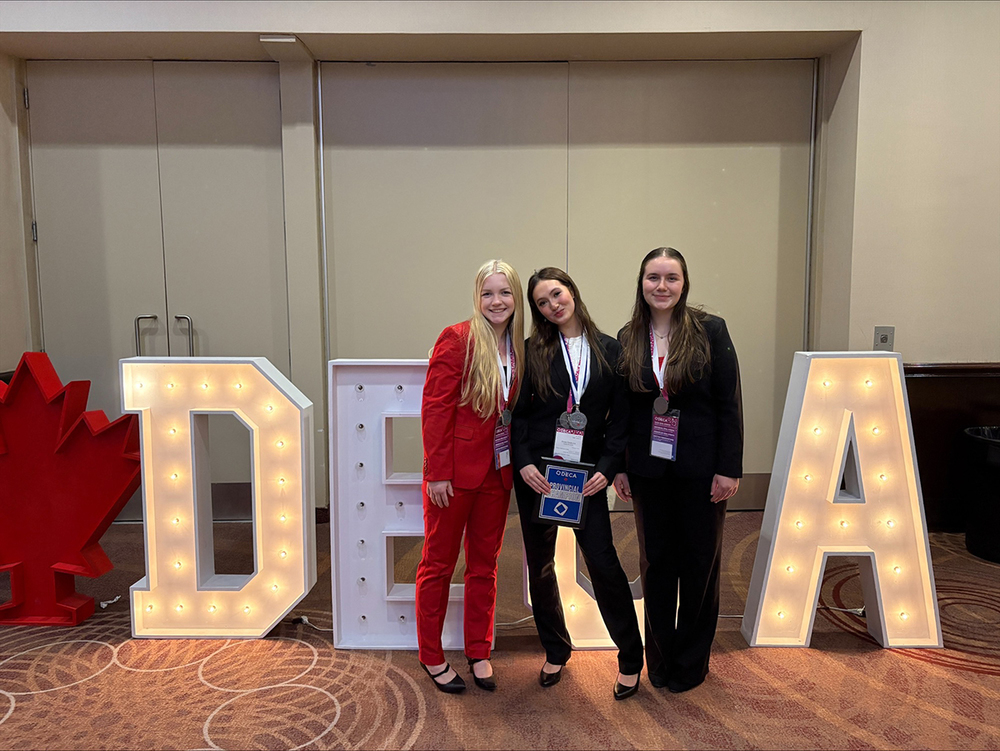 3 students pose for a photo in front of a DECA sign.