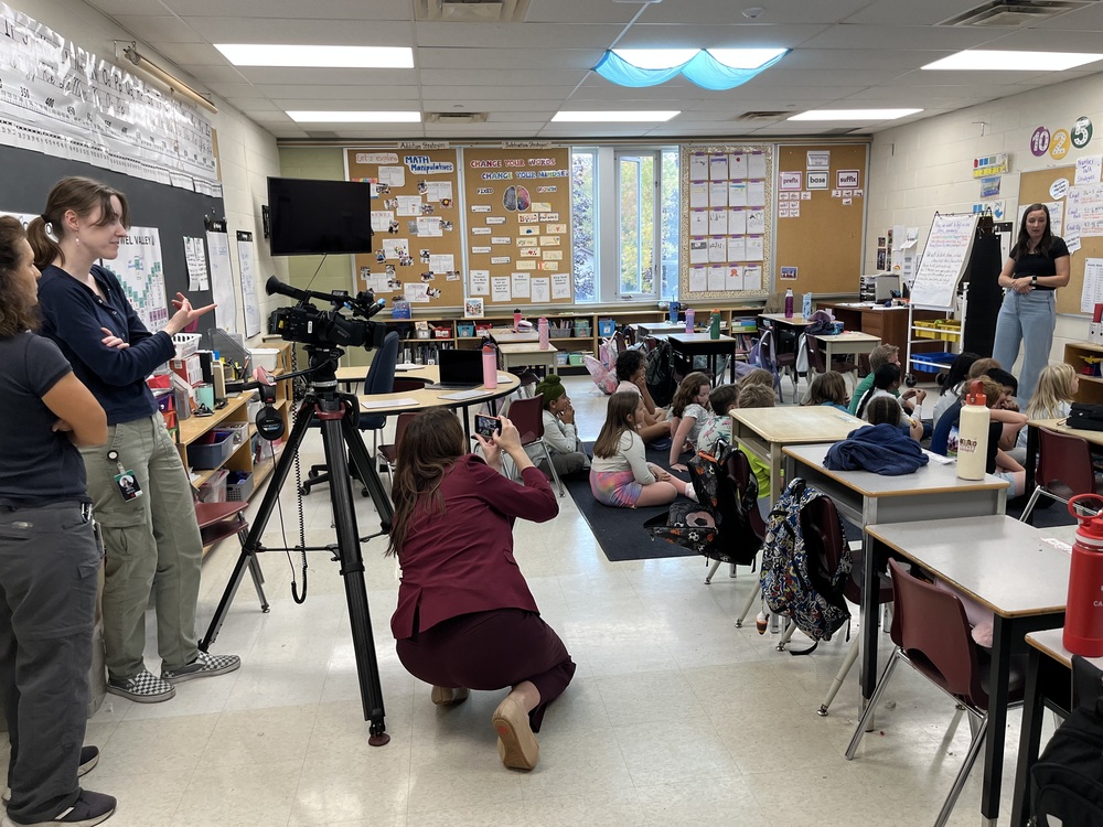 Report kneeling and taking photos of children learning on carpet, while teacher is teaching a math lesson. Camera and lights behind photographer. 