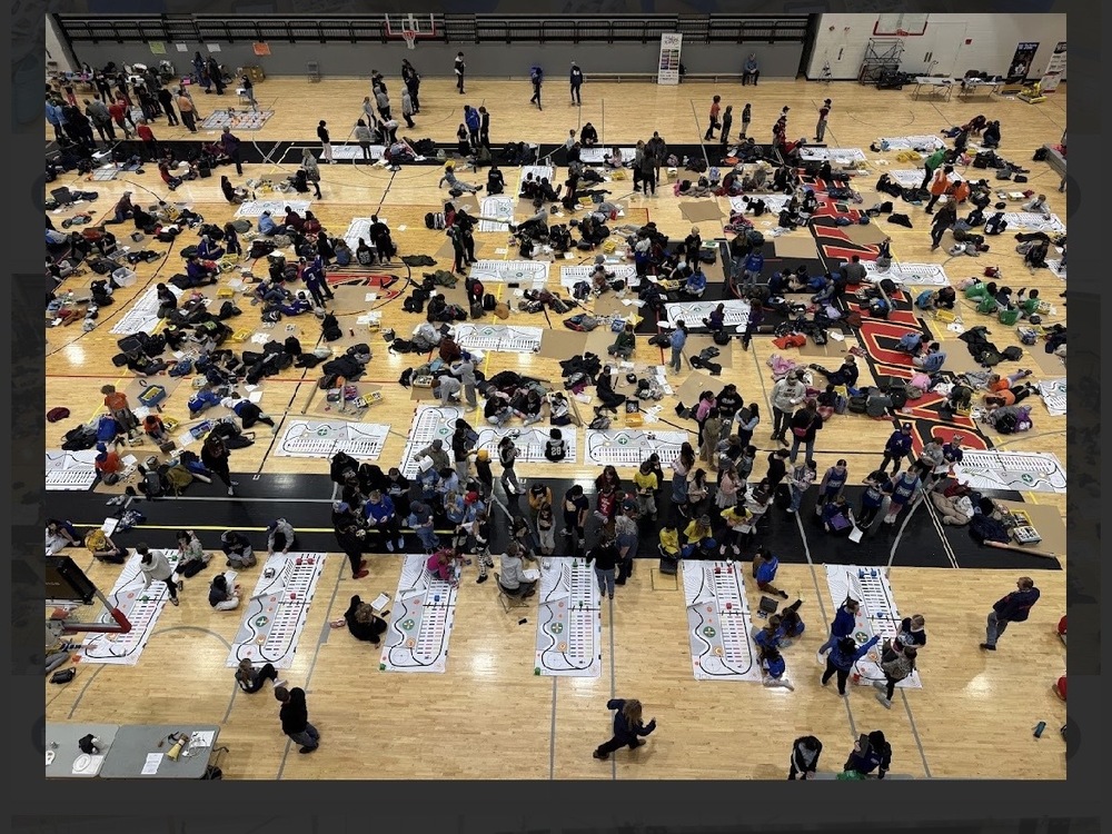 Gym floor full of students working on coding tasks. There are white boards with various routes. 