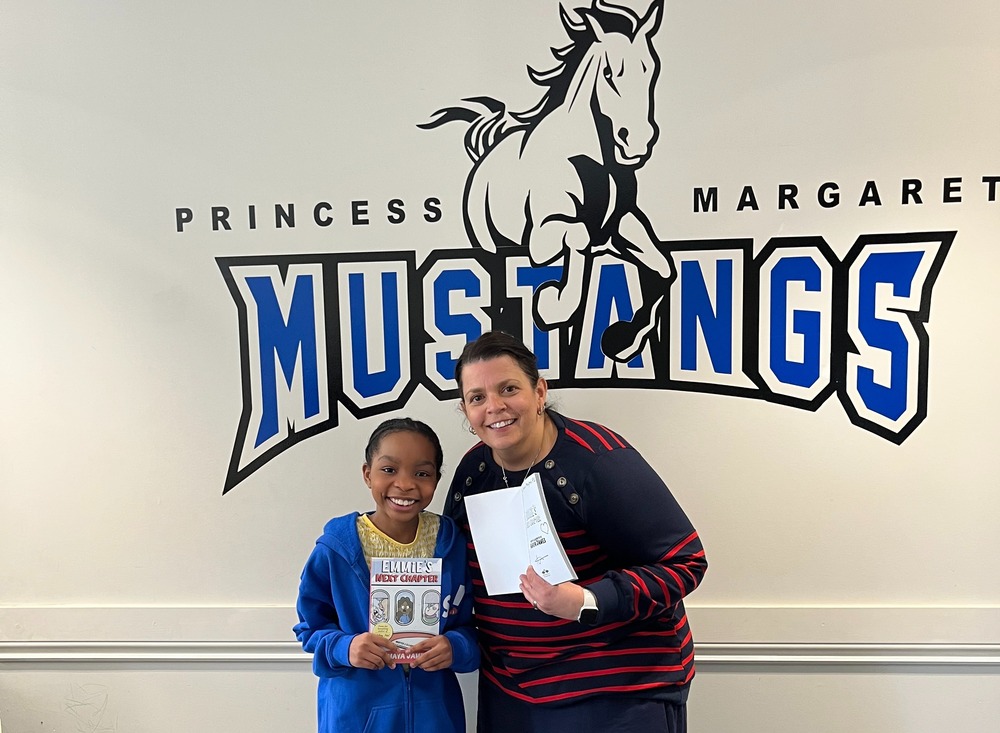 Standing in our front of our blue and black school logo ( PMPS Mustangs) with student and principal holding copies of the students new book.