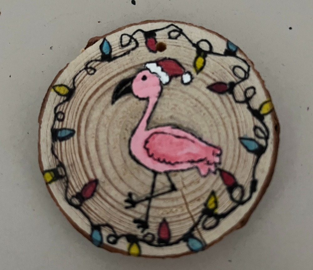 A painted pink flamingo on a round piece of wood with Christmas lights around it and a Santa hat on he bird. 