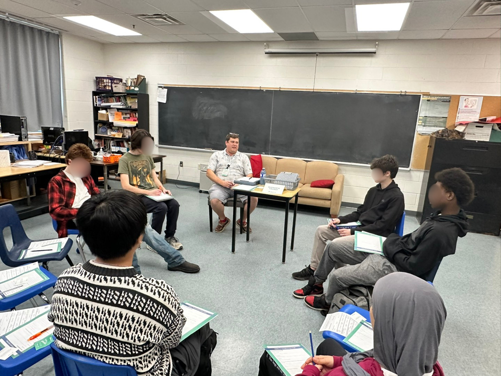 A group of people sit in a circle in chairs inside a classroom.