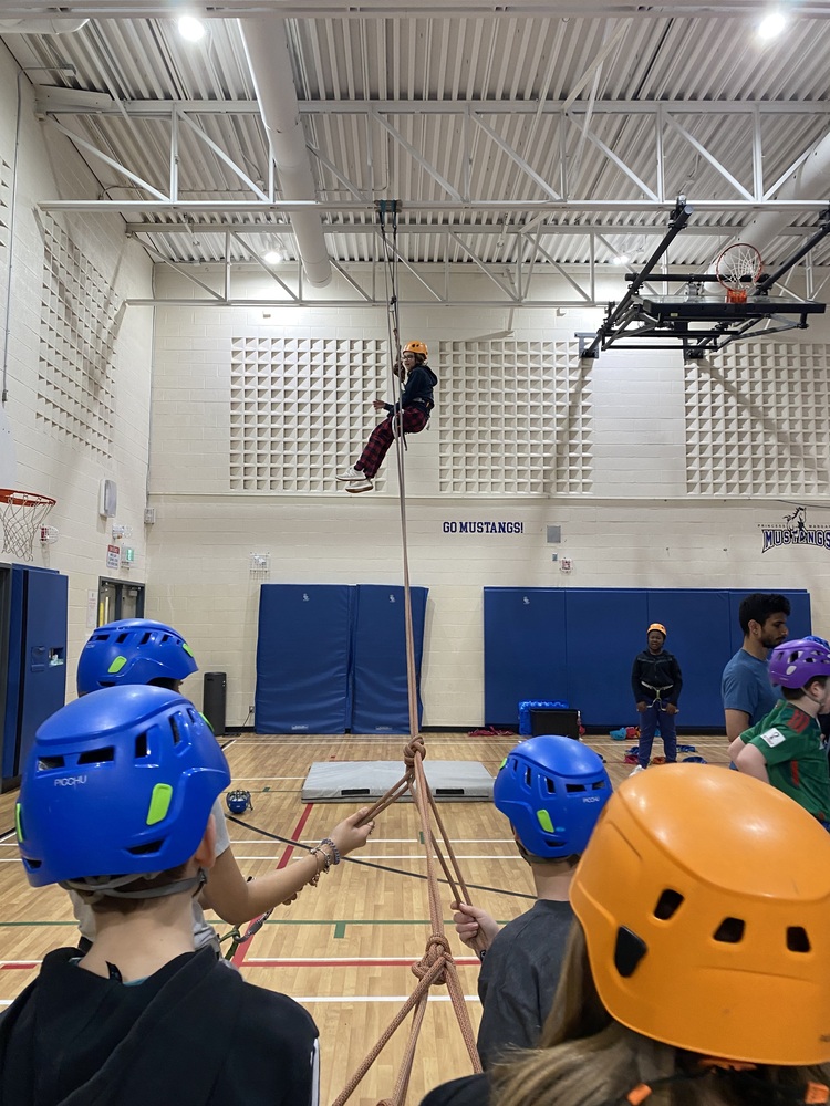 Children with helmets and harnesses pulling another child to the top of the gym ceiling. 