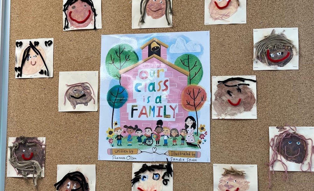 Class board with a picture of a book cover "Our Class is a Family" with self portraits of children with yarn, paint, buttons etc.
