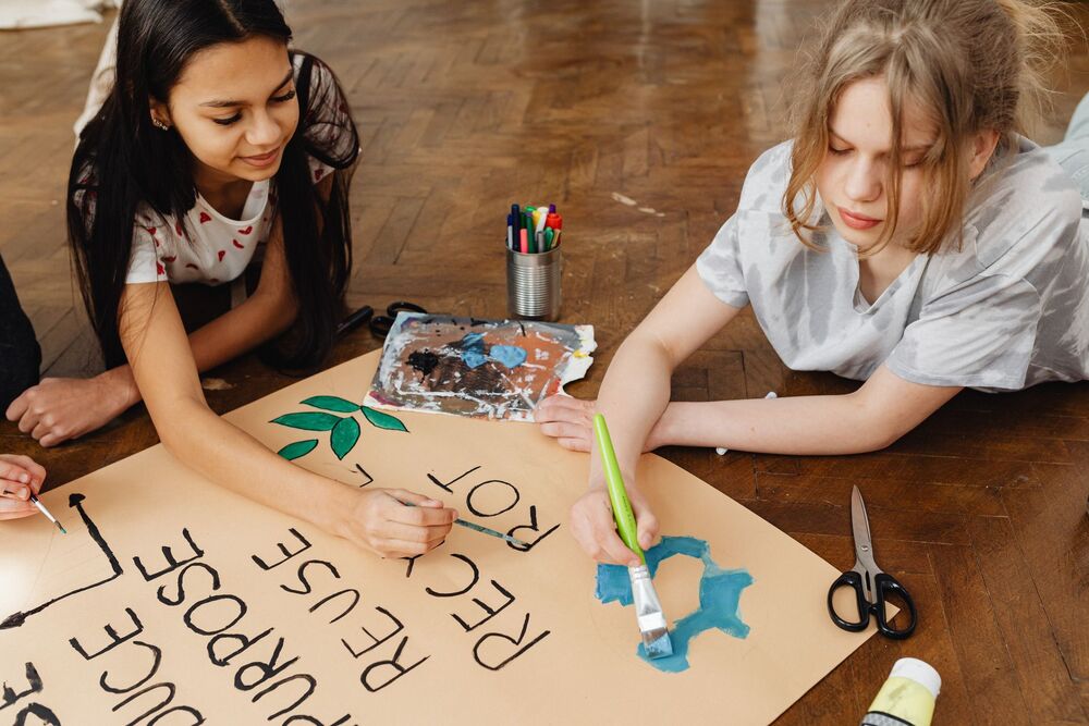 Two children write on a poster with words like 'recycle, reuse, repurpose'