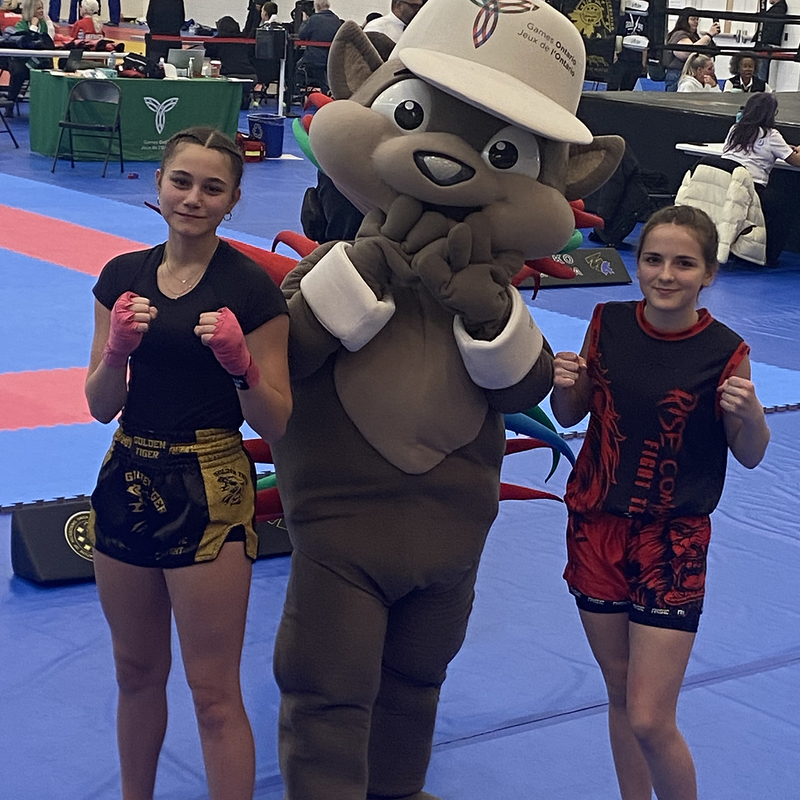 2 kickboxers stand on a competition floor with a mascot from the Ontario Winter Games