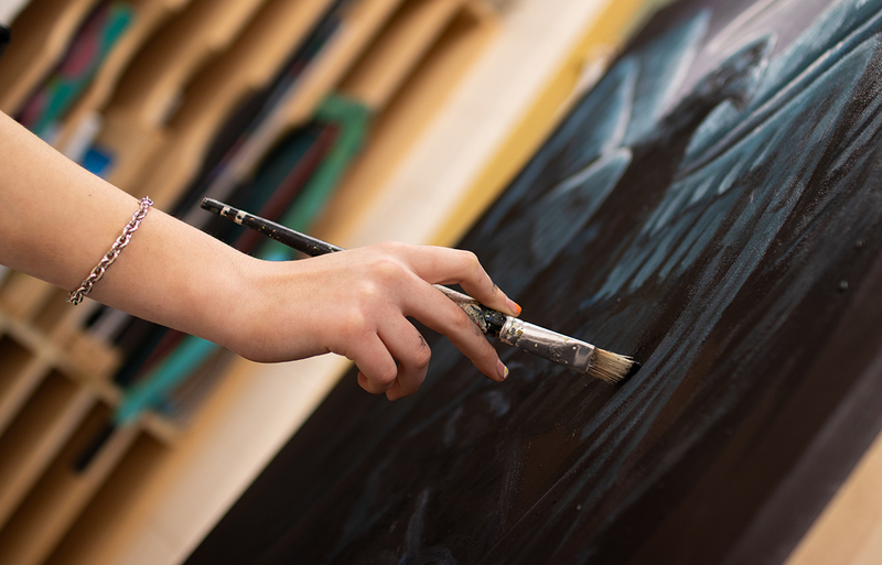 Photograph of a person's hand holding a paint brush and applying paint to a canvas