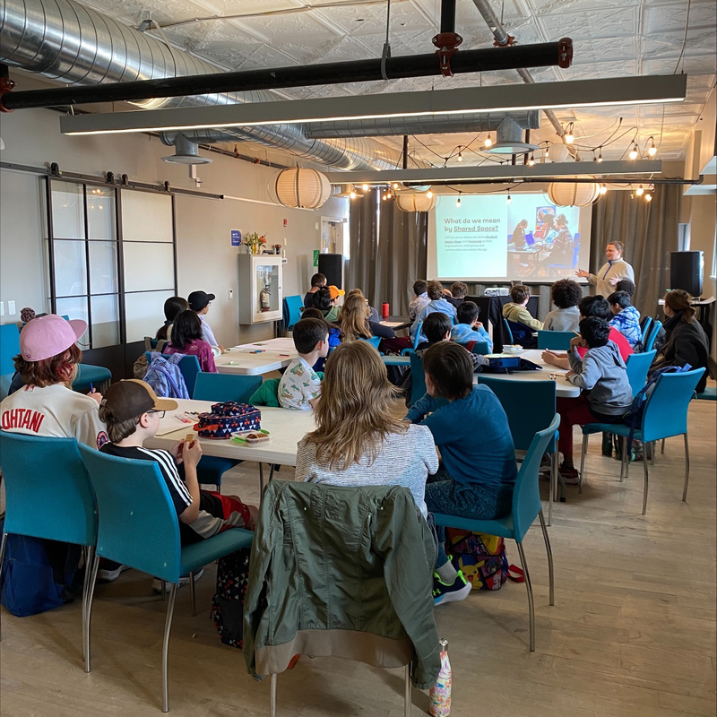 Students sit at tables in a large room watching a presentation.